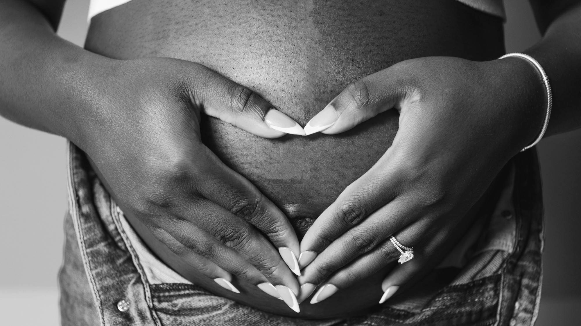 Black-and-white close-up of two hands forming a heart over a pregnant belly.